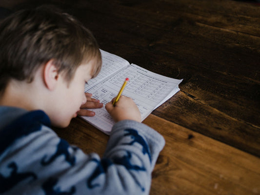 A young boy with short light brown hair, wearing a blue sea-creature patterned sweater, sits at a rustic wooden table and concentrates on writing in a notebook with a yellow pencil. The scene feels warm and homey, with the child focused on his worksheet.